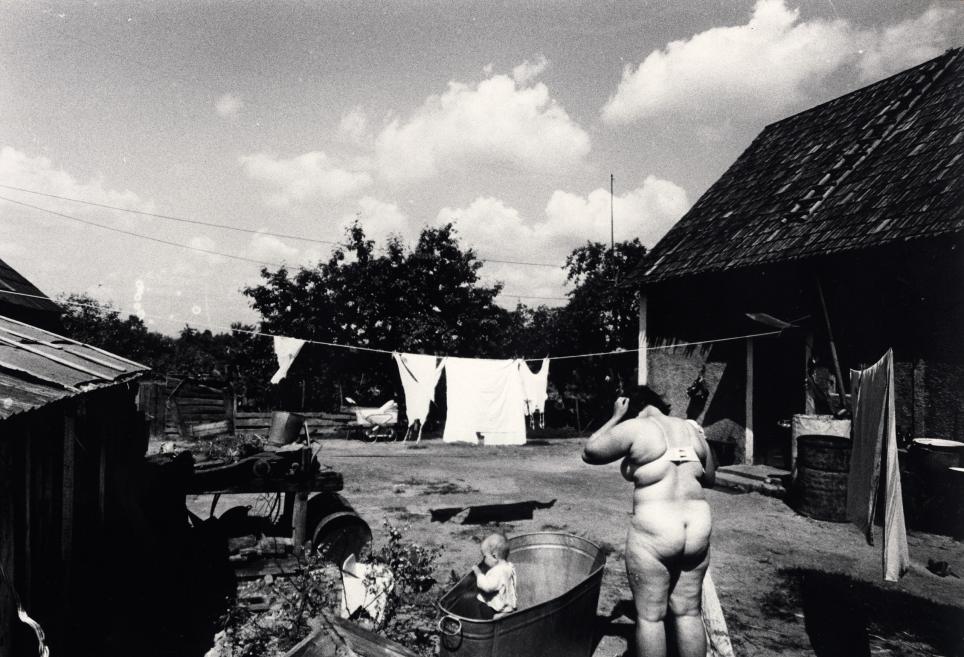 Melnbalta fotogrāfija ar sievieti peldkostīmā pie vannas pagalmā starp mājām, veļu auklām un bērnu vannā.
