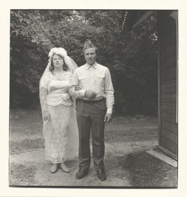 Melnbalta kāzu fotogrāfija ar līgavu baltā mežģīņu kleitā un plīvurā un līgavaini gaišā kreklā, stāvot ārā pie koka ēkas un apstādījumu fona.