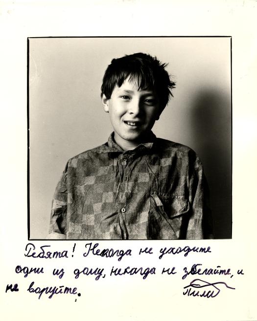 Melnbalta portreta fotogrāfija ar zēnu rūtainā krekliņā uz vienkārša fona, zem attēla ar roku rakstīts krievu valodas teksts.