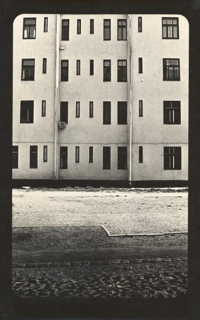 Black-and-white photograph of an apartment building facade with multiple windows, seen from a street with cobblestones in the foreground.