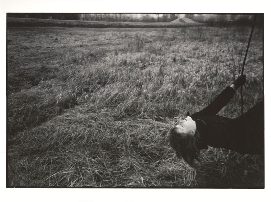 Black-and-white photograph showing a woman lying in grass and holding a curved rod or rope in an open field.