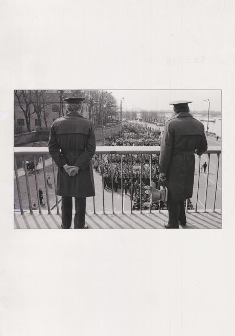 Black-and-white photograph of two uniformed men standing by a railing and looking down at a large street procession beside a river.