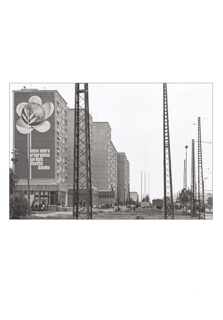 Black-and-white cityscape with an apartment building bearing a large mural of a flower and globe with text, and several tall metal poles in the foreground.