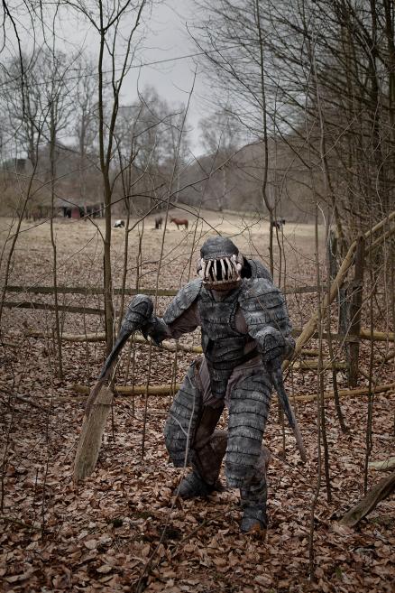 A figurative costume or sculptural figure in dark armor stands in a wooded, leaf-covered field with bare trees and horses visible in the distance.
