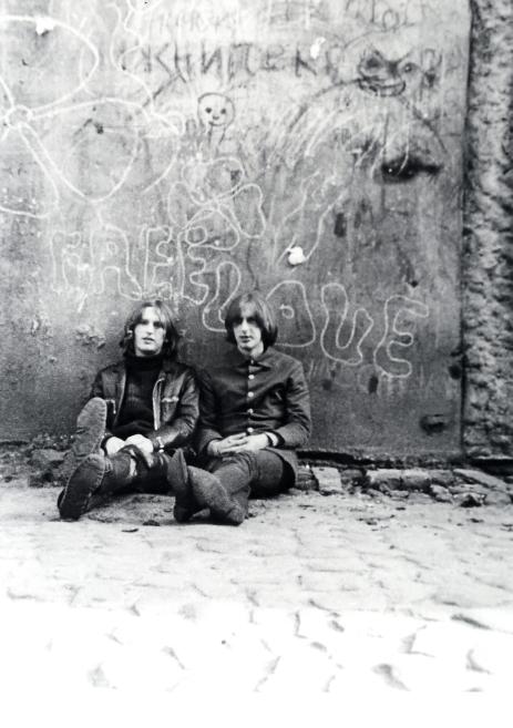 Black-and-white photograph of two long-haired young men sitting on cobblestones in front of a wall covered with graffiti.