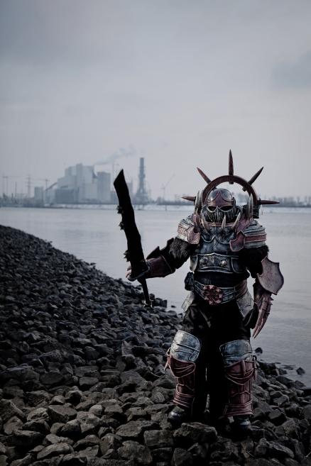 An armed figure in fantasy armor stands on a rocky shoreline by the water, with an industrial landscape in the misty background.