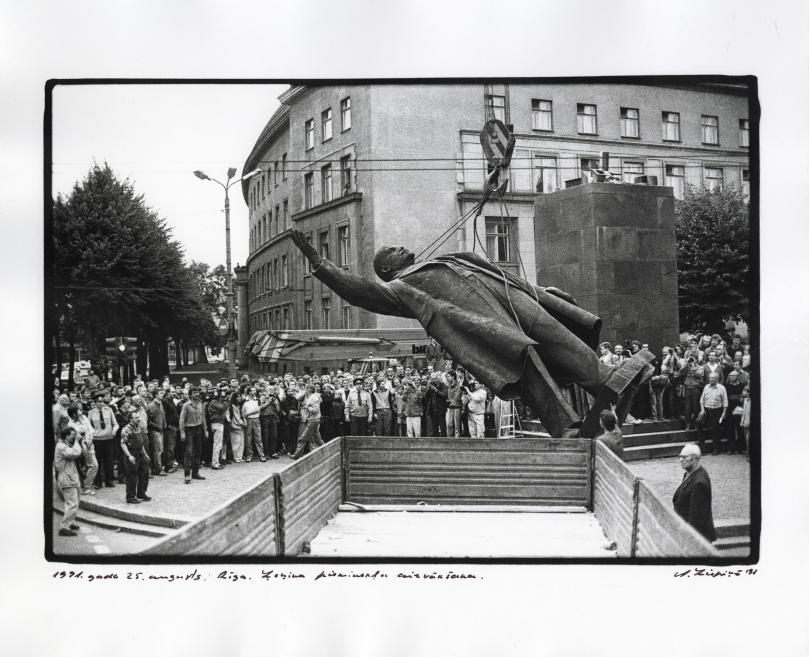 Black-and-white photograph of a reclining male statue suspended by crane cables in a city square, surrounded by a large crowd of onlookers.