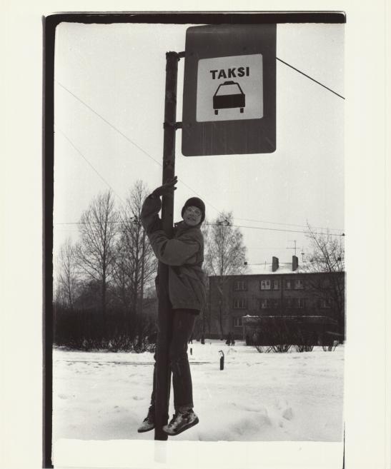 Black-and-white photograph of a person in winter clothing clinging to a pole beside a street taxi sign marked TAKSI with a car symbol, in a snowy setting.