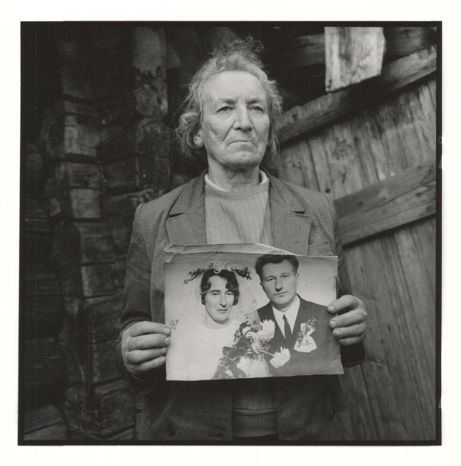 Black-and-white portrait of an elderly woman standing by a wooden wall and holding a wedding photograph.