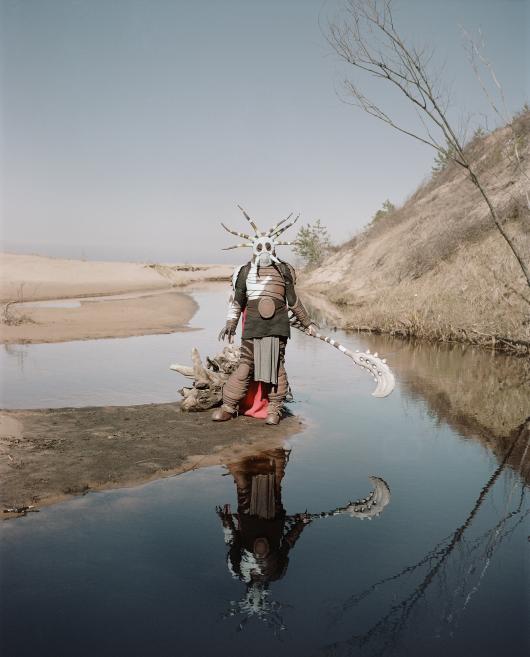 A person in a fantasy costume with a mask and spear stands by a small river in a sandy landscape, reflected in the water.