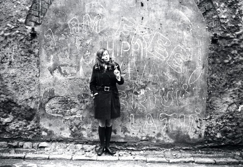 In a black-and-white photograph, a woman stands by a graffiti-covered wall holding a cigarette.