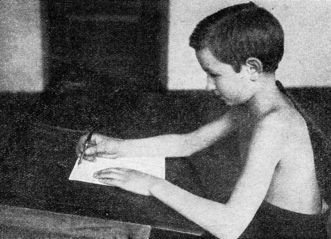 A boy writing on a white sheet at a school desk.