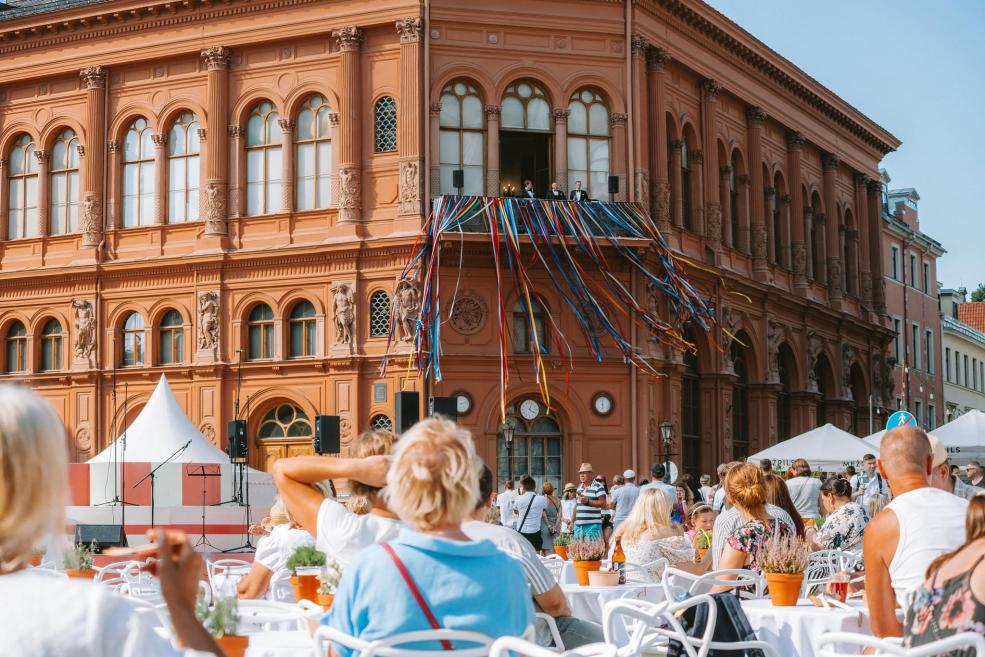 Latvijas Nacionālā mākslas muzeja ēkas fasāde ar krāsainām lentēm pie balkona un cilvēku pūli āra pasākumā priekšplānā.