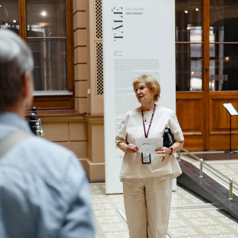 Muzeja vestibilā sieviete vada ekskursiju apmeklētāju grupai pie izstādes informatīvā stenda.