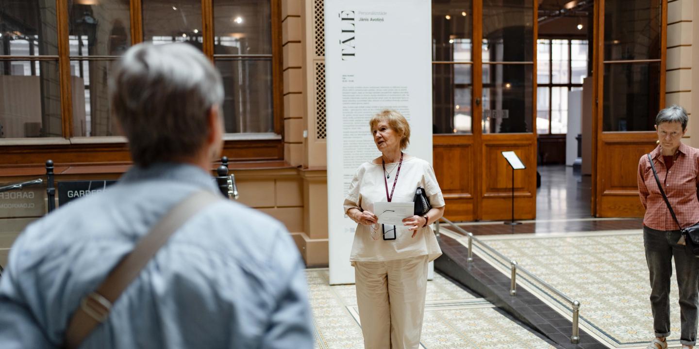 Muzeja vestibilā sieviete vada ekskursiju apmeklētāju grupai pie izstādes informatīvā stenda.