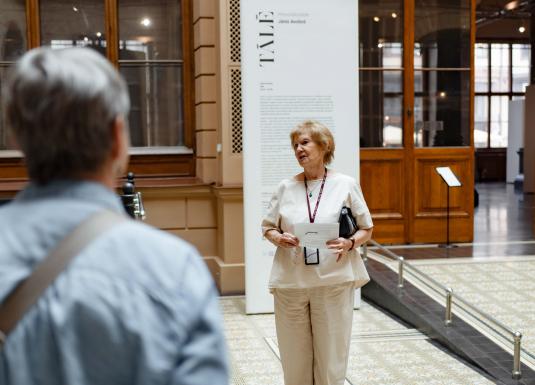 Muzeja vestibilā sieviete vada ekskursiju apmeklētāju grupai pie izstādes informatīvā stenda.