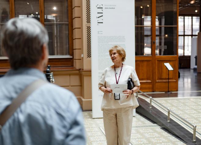 Muzeja vestibilā sieviete vada ekskursiju apmeklētāju grupai pie izstādes informatīvā stenda.