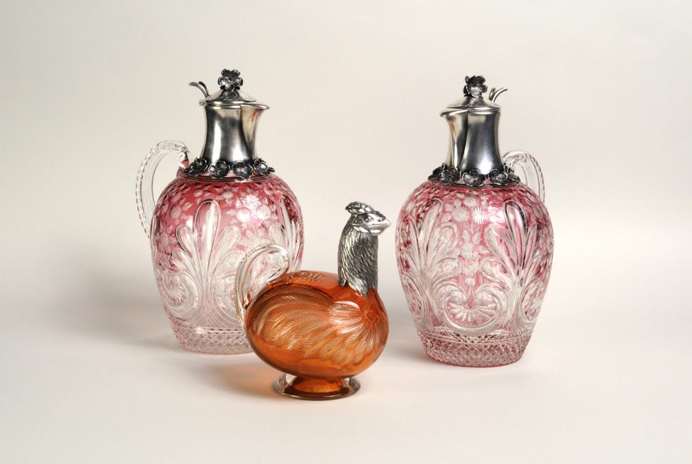 Two pink engraved glass decanters with metal lids and an orange glass bird-shaped vessel on a light background.