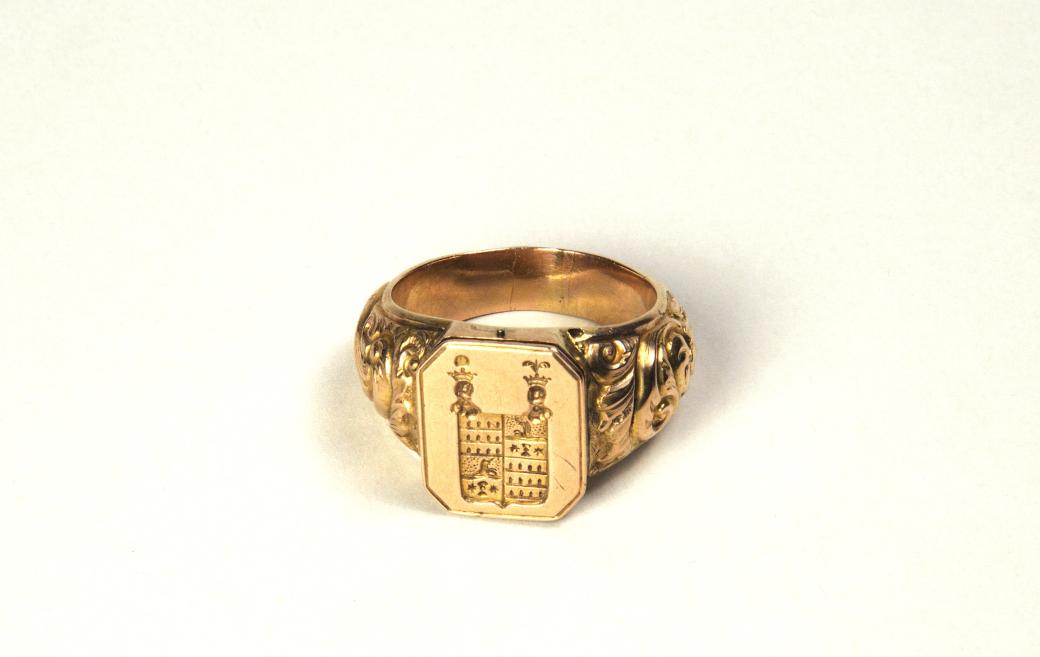 Gold ring with an engraved coat of arms and decorative ornaments on a light background.