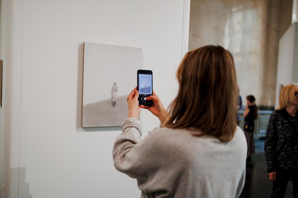 Apmeklētāja ar viedtālruni fotografē neliela formāta gleznu, kas izvietota uz baltas ekspozīcijas sienas muzeja izstāžu zālē.