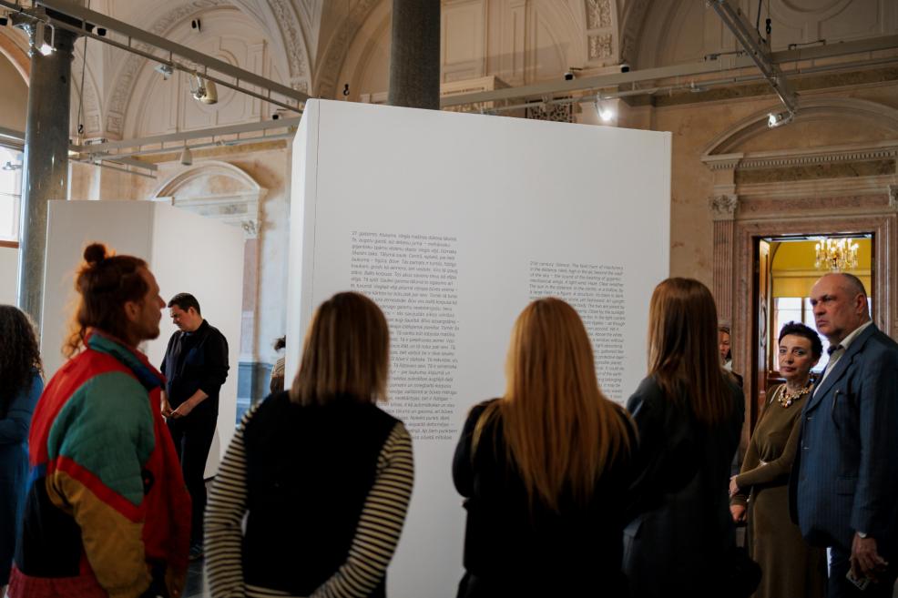 Apmeklētāji pulcējas pie ekspozīcijas sienas ar izstādes tekstu, lasot informāciju un apspriežot redzēto muzeja vēsturiskajā izstāžu zālē.