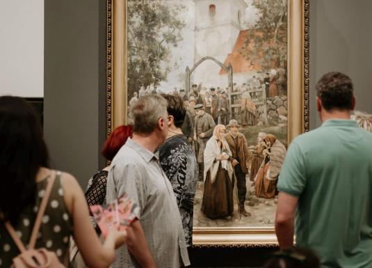 Muzeja apmeklētāji pulcējušies pie lielas figurālas gleznas, kurā attēlota cilvēku grupa baznīcas pagalmā.