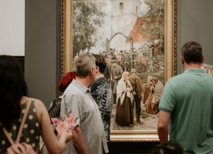 Muzeja apmeklētāji pulcējušies pie lielas figurālas gleznas, kurā attēlota cilvēku grupa baznīcas pagalmā.