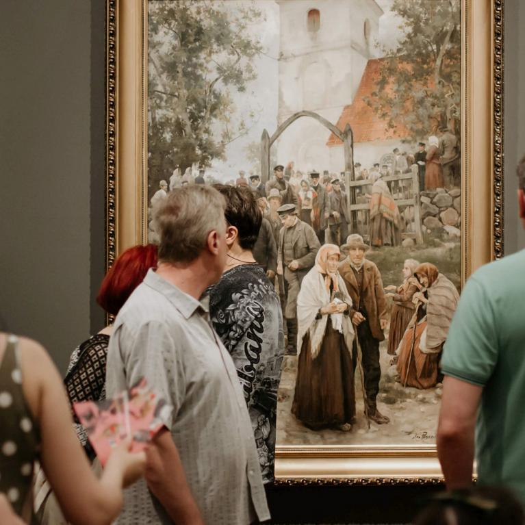 Muzeja apmeklētāji pulcējušies pie lielas figurālas gleznas, kurā attēlota cilvēku grupa baznīcas pagalmā.