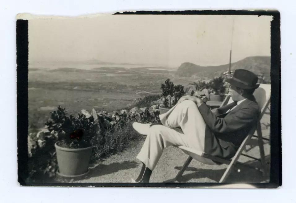 Vēsturiska melnbalta fotogrāfija attēlo cilvēku cepurē, sēžot atpūtas krēslā un raugoties plašā ainavā no terases.