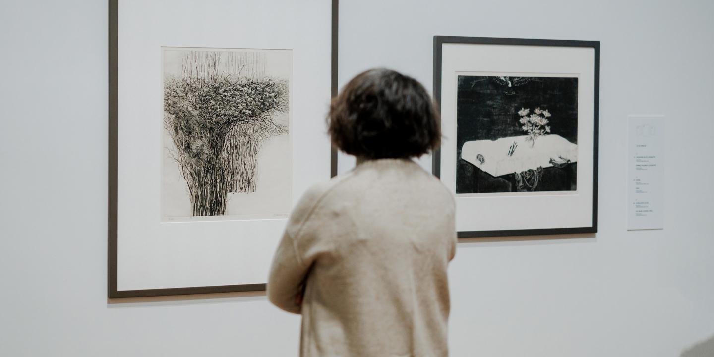Apmeklētājs muzejā aplūko divus ierāmētus grafikas darbus uz baltas sienas.