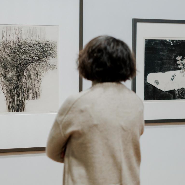 Apmeklētājs muzejā aplūko divus ierāmētus grafikas darbus uz baltas sienas.