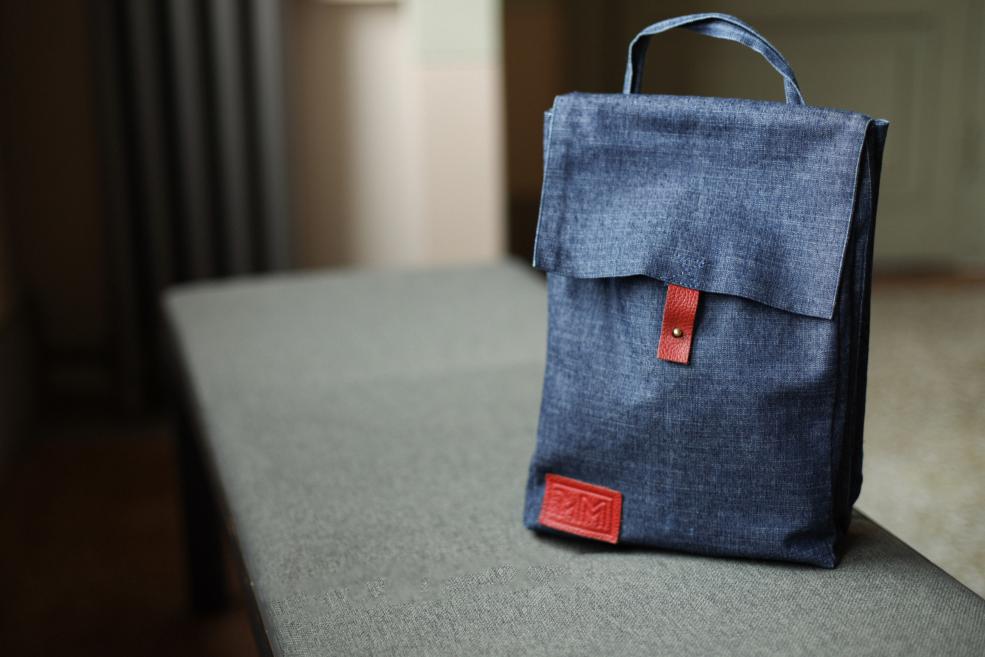 Tumši zila auduma soma ar sarkanas ādas detaļām uz pelēka galda.