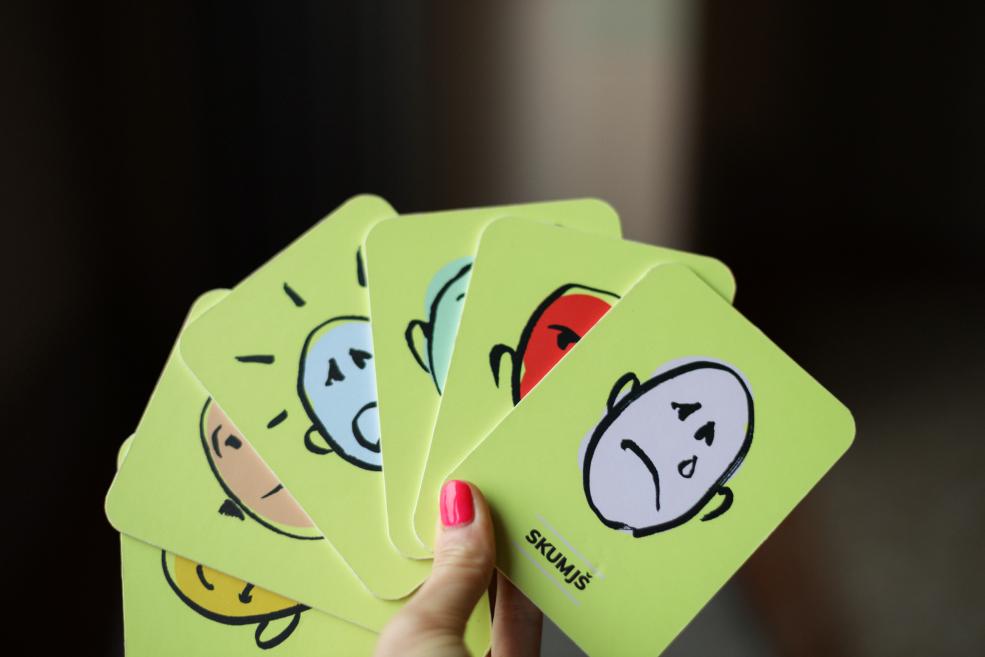 Rokā turētas emociju kartītes ar dažādām sejas izteiksmēm uz zaļa fona.