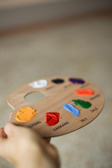 Rokā turēta koka krāsu palete ar dažādu toņu akrila krāsām un uzrakstiem latviešu valodā.