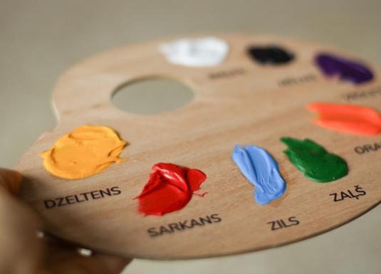 Koka krāsu palete ar uzziestām akrila krāsām un nosaukumiem latviešu valodā – dzeltena, sarkana, zila, zaļa u.c.