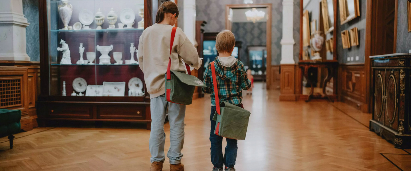 Two children with shoulder bags walk through a museum hall displaying porcelain dishes and paintings in a classical interior.
