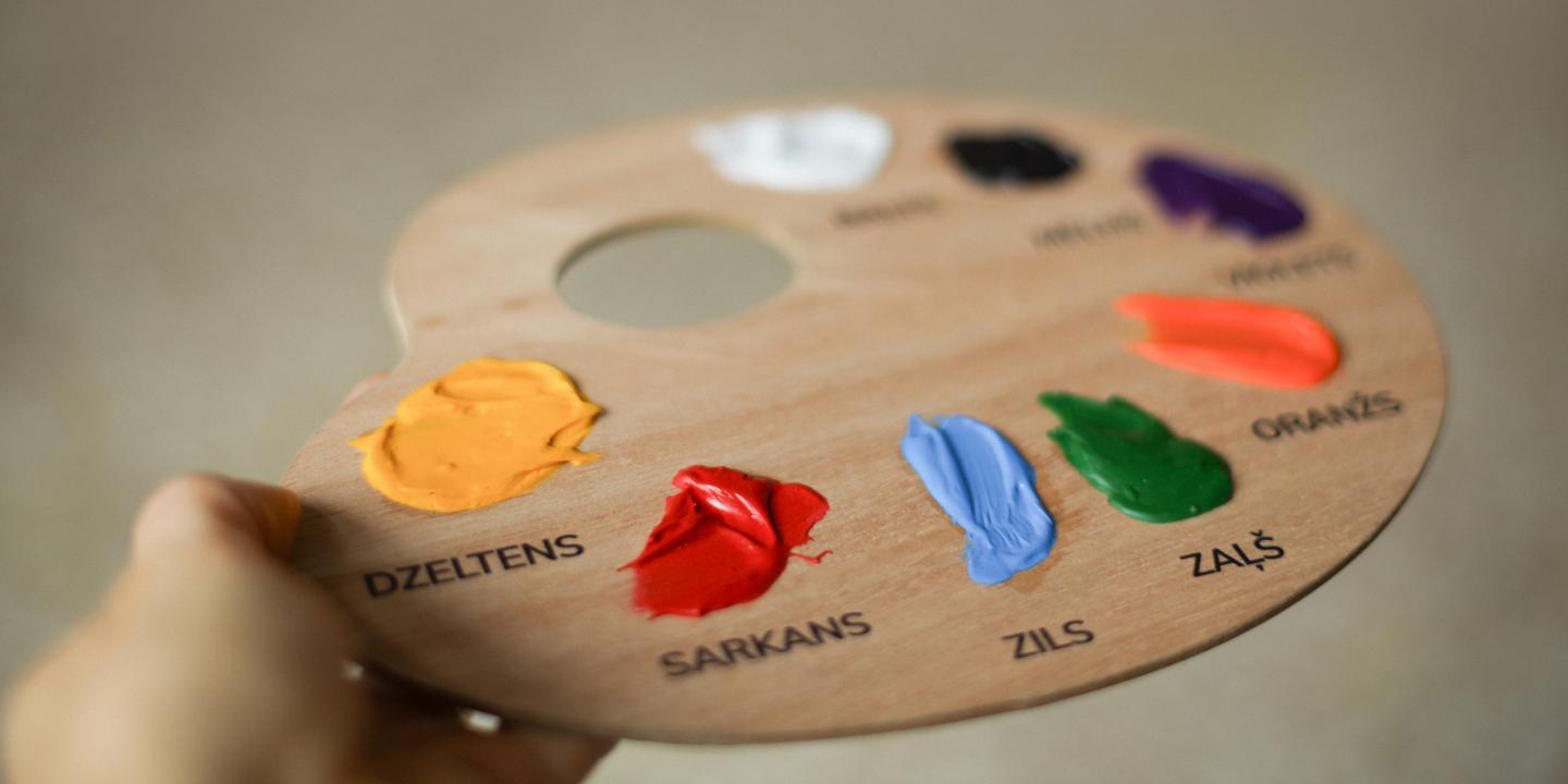 Koka krāsu palete ar uzziestām akrila krāsām un nosaukumiem latviešu valodā – dzeltena, sarkana, zila, zaļa u.c.