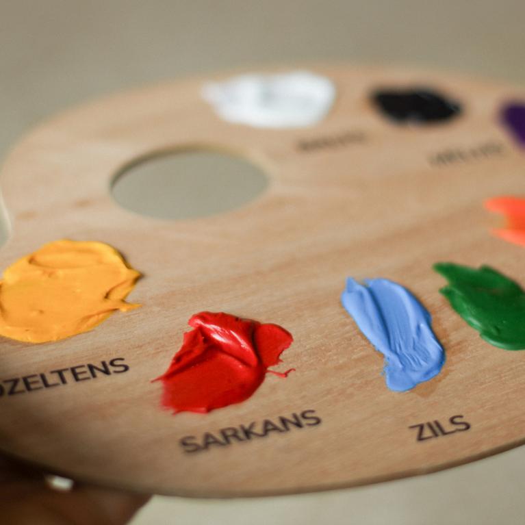 Koka krāsu palete ar uzziestām akrila krāsām un nosaukumiem latviešu valodā – dzeltena, sarkana, zila, zaļa u.c.