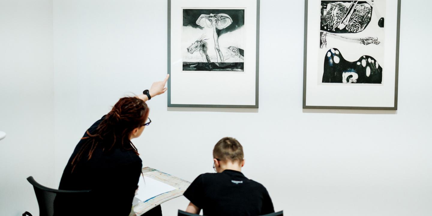 Sieviete muzejā rāda uz grafikas darbu pie sienas, kamēr zēns sēž un zīmē, skatoties uz attēlu.