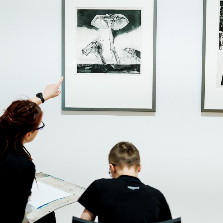 Sieviete muzejā rāda uz grafikas darbu pie sienas, kamēr zēns sēž un zīmē, skatoties uz attēlu.