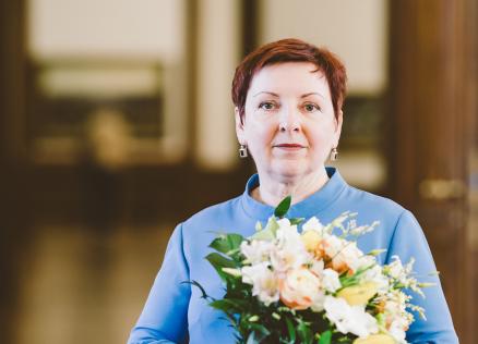 Sieviete ar īsiem matiem stāv telpā un tur rokās gaišu ziedu pušķi, skatoties kamerā.