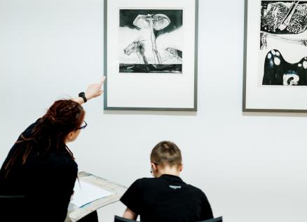 Sieviete muzejā rāda uz grafikas darbu pie sienas, kamēr zēns sēž un zīmē, skatoties uz attēlu.