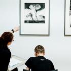 Sieviete muzejā rāda uz grafikas darbu pie sienas, kamēr zēns sēž un zīmē, skatoties uz attēlu.