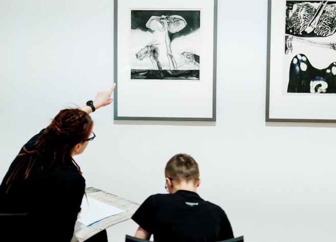 Sieviete muzejā rāda uz grafikas darbu pie sienas, kamēr zēns sēž un zīmē, skatoties uz attēlu.