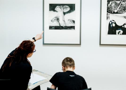 Sieviete muzejā rāda uz grafikas darbu pie sienas, kamēr zēns sēž un zīmē, skatoties uz attēlu.