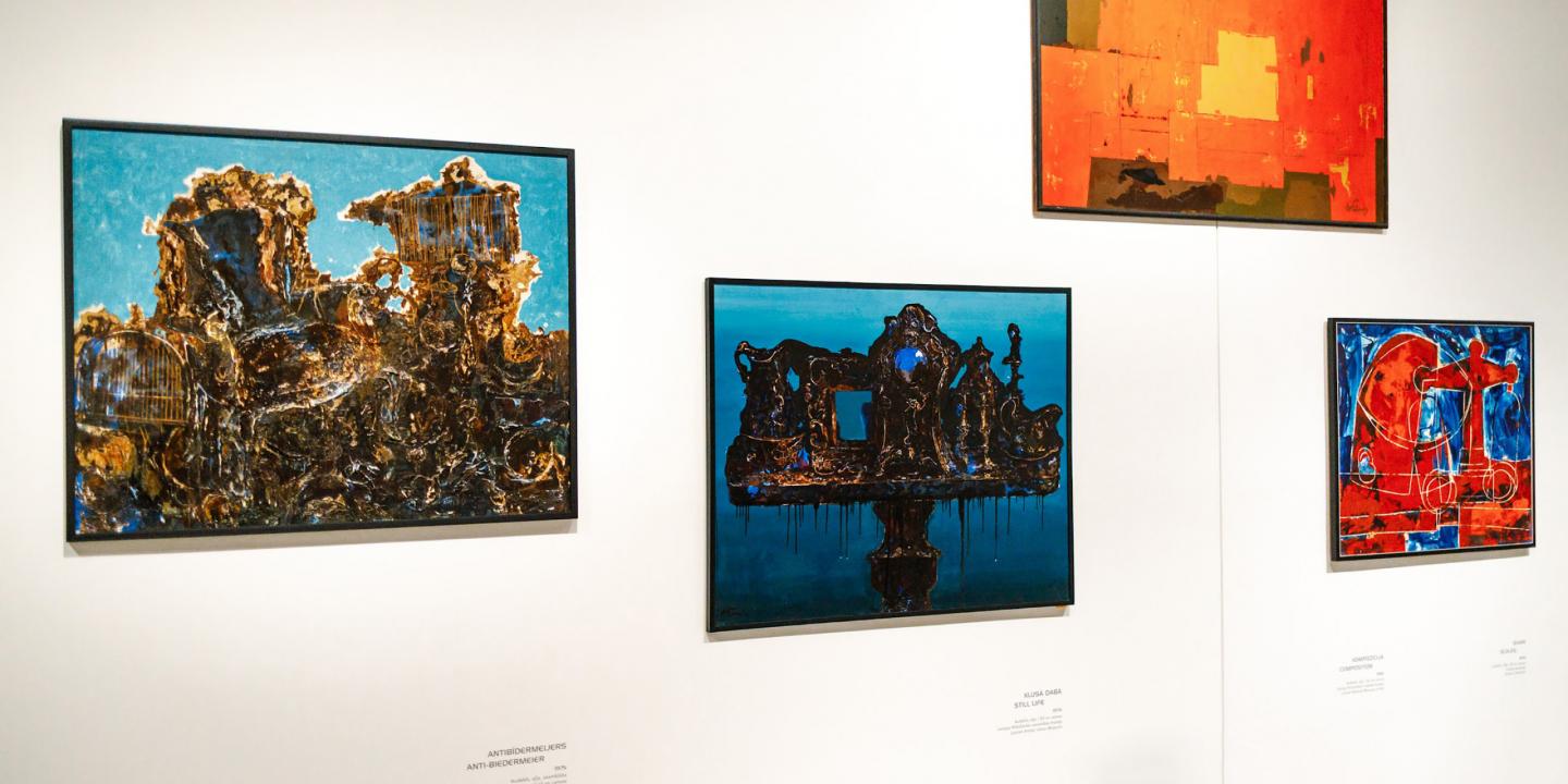 Muzeja sienā izkārtotas četras krāsainas abstraktas gleznas dažādos formātos ar izteiktiem zilas, sarkanas un zeltainas krāsas laukumiem.