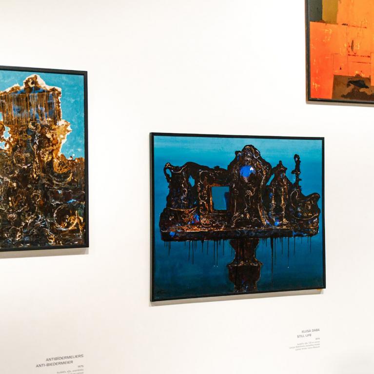 Muzeja sienā izkārtotas četras krāsainas abstraktas gleznas dažādos formātos ar izteiktiem zilas, sarkanas un zeltainas krāsas laukumiem.