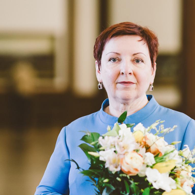 Sieviete ar īsiem matiem stāv telpā un tur rokās gaišu ziedu pušķi, skatoties kamerā.