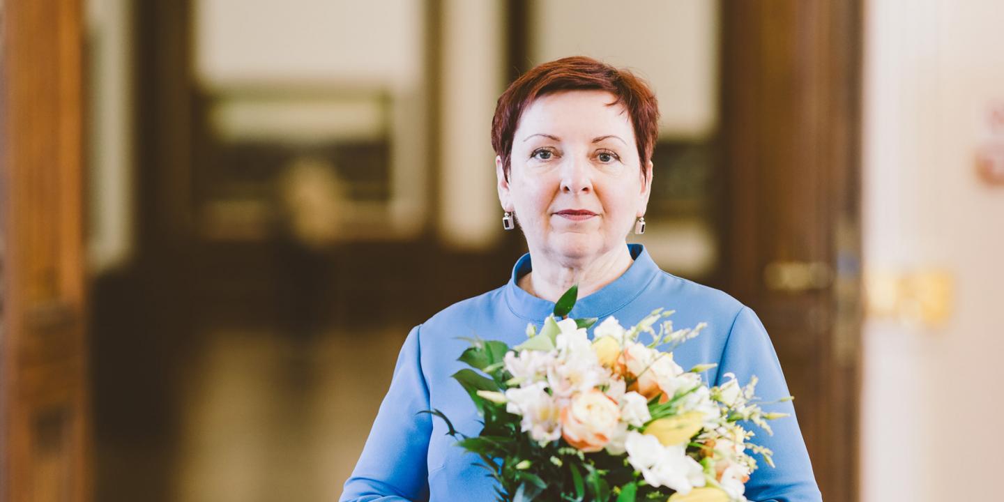 Sieviete ar īsiem matiem stāv telpā un tur rokās gaišu ziedu pušķi, skatoties kamerā.