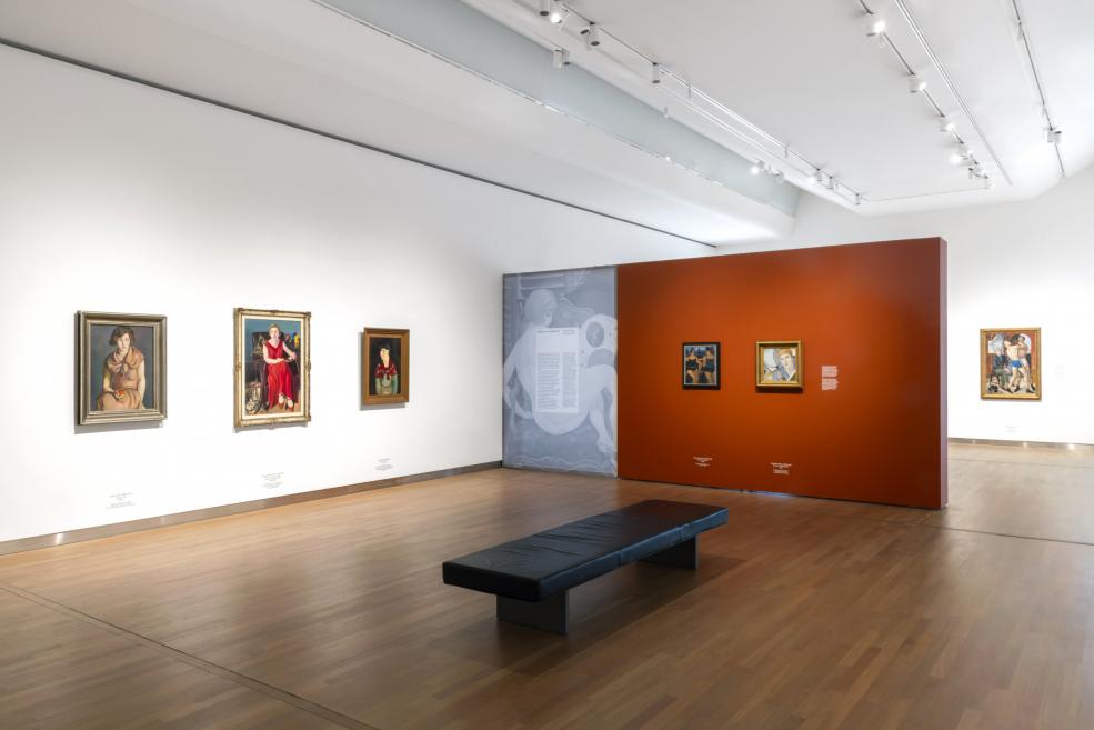 Izstāžu zālē ar modernisma portretiem un centrā novietotu soliņu skatāmas gleznas uz baltas un oranžas sienas.